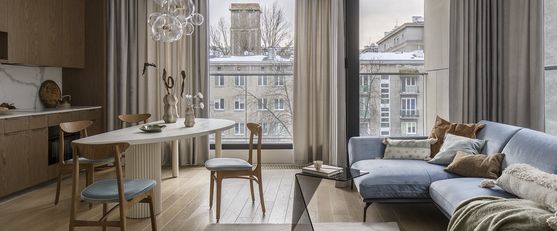 Stylowe mieszkanie z otwartą kuchnią i salonem, wyposażone w drewniane meble, jasną sofę w kolorze niebieskim, owalny stół z dekoracyjnymi wazonami oraz duże okna z widokiem na miejską architekturę.